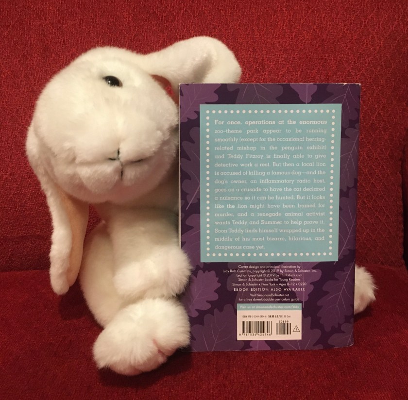 Marshmallow is pointing to the back cover of the fifth book, Lion Down, in the FunJungle series by Stuart Gibbs, where the reader can get a quick summary of the plot of the book.
