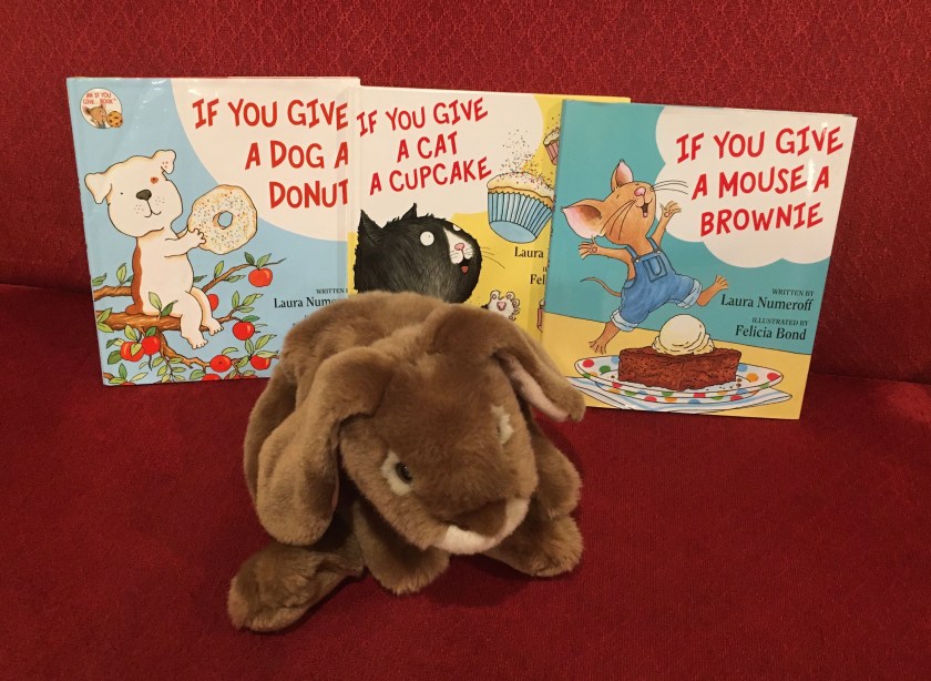Caramel is showing the readers If You Give a Cat a Cupcake (2008), If You Give a Dog a Donut (2011), and If You Give a Mouse a Brownie (2016), all written by Laura Numeroff and illustrated by Felicia Bond.