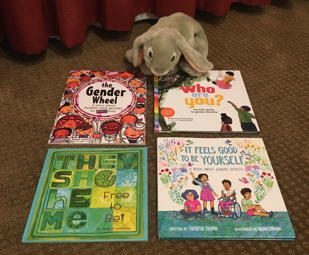 Sprinkles is posing with four books that explain pronouns and gender identity to children: The Gender Wheel by Maya Gonzalez, Who Are You? The Kid's Guide to Gender Identity, written by Brook Pessin-Whedbee and illustrated by Naomi Bardoff, It Feels Good To Be Yourself, written by Theresa Thorn and illustrated by Noah Grigni, and They, She, He, Me: Free To Be! by Maya Gonzalez and Matthew Smith-Gonzalez. 