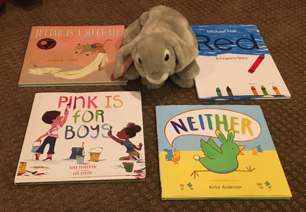Sprinkles is posing with four books that emphasize that being different is okay: Julián Is A Mermaid by Jessica Love, Red: A Crayon's Story by Michael Hall, Neither by Airlie Anderson, and Pink Is For Boys written by Robb Pearlman and illustrated by Eda Kaban.  