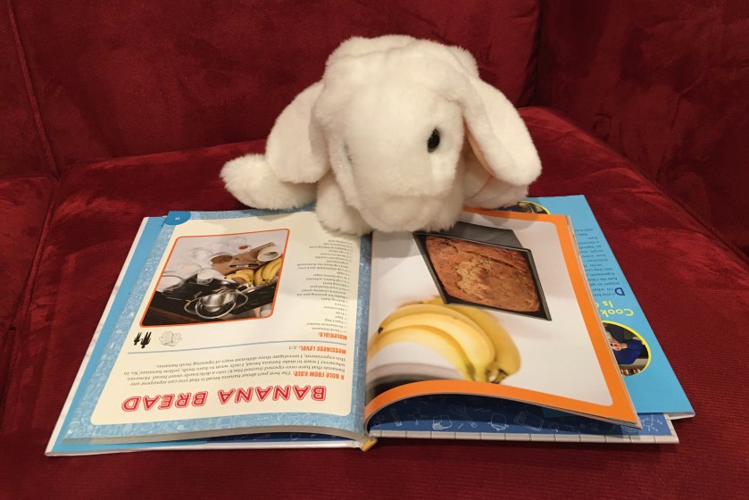 Marshmallow is reading about banana bread in The Awesome Book of Edible Experiments for Kids by Kate Biberdorf. 